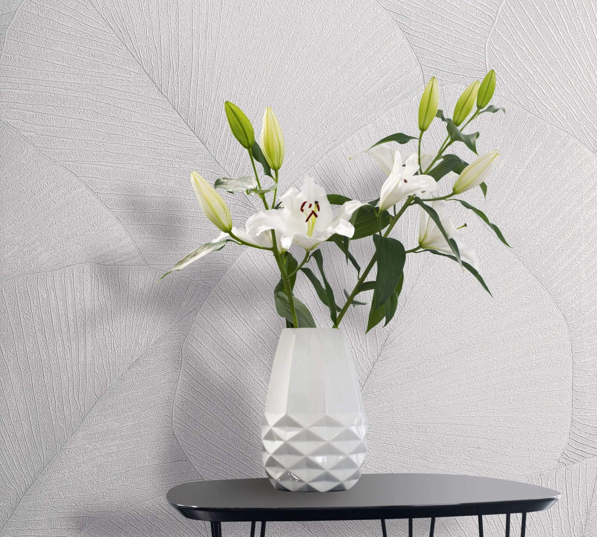 Marburg Kumano 34507 White wallpaper with large overlapping leaf vein pattern, shown behind a faceted white vase of lilies on a small black table.