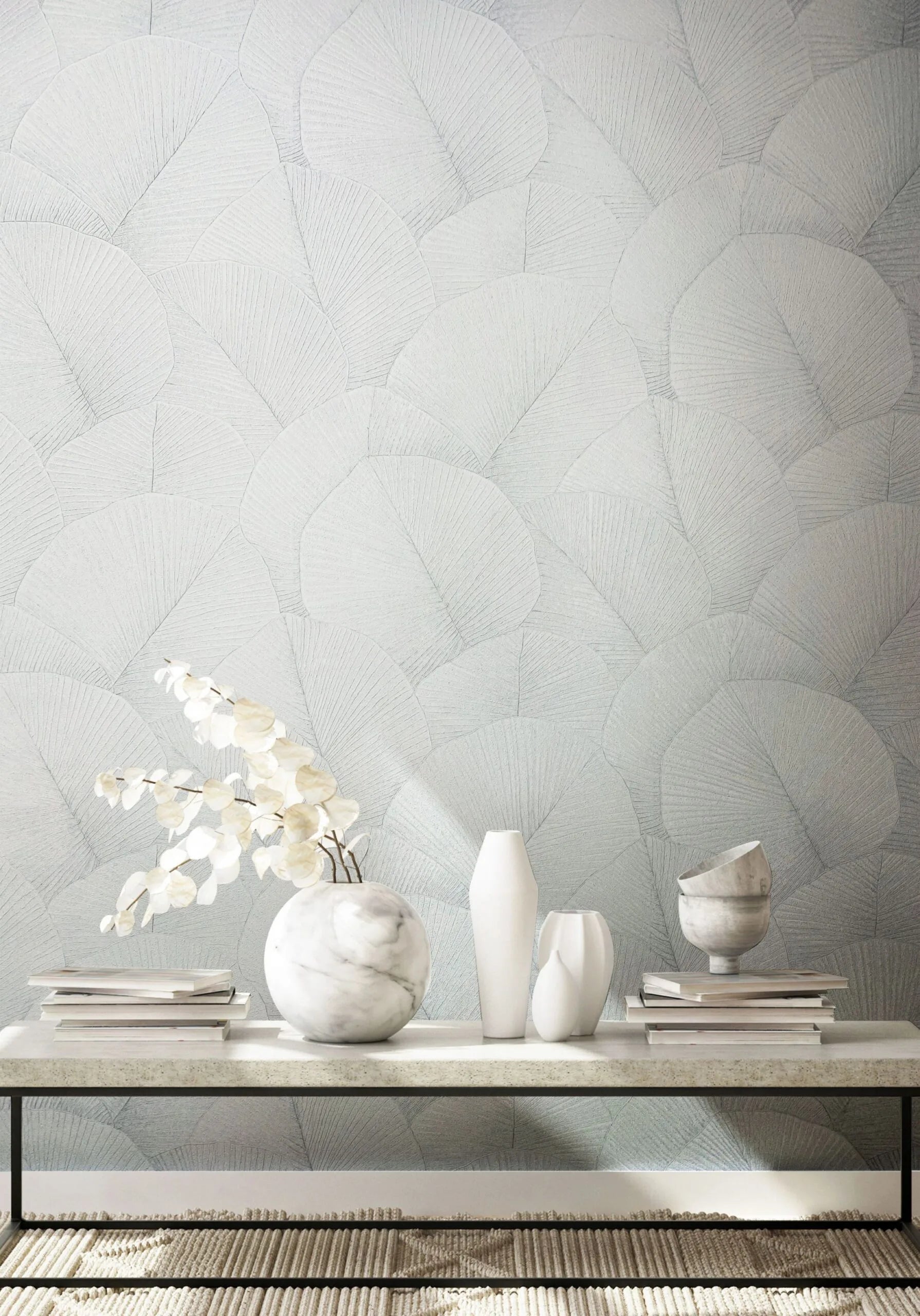 Marburg Kumano 34507 Light neutral wallpaper with overlapping fan-like leaf pattern behind a low bench styled with white and marble vases, stacked books, and a small branch; woven rug visible at the bottom.