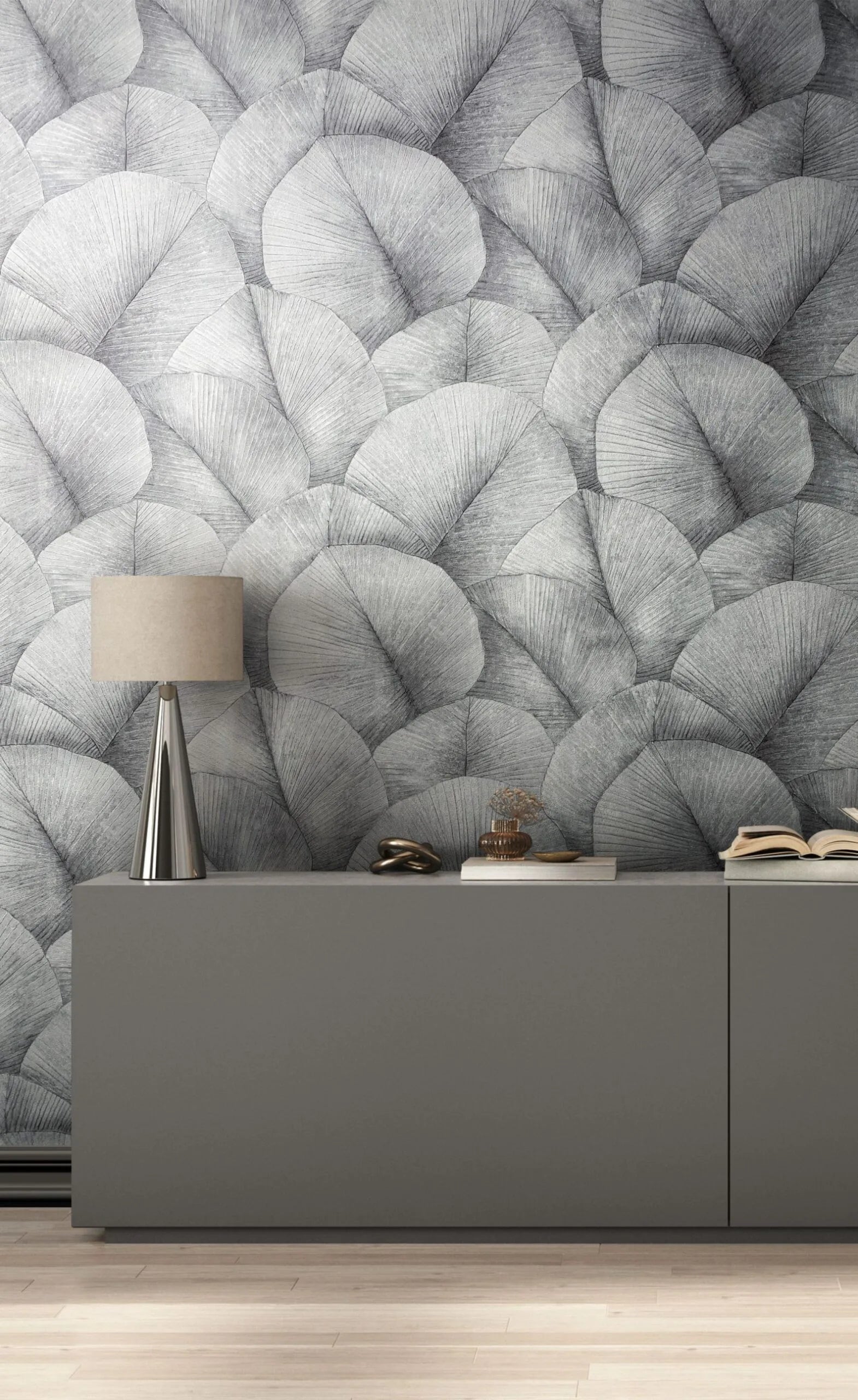 Marburg Kumano 34508 Large-scale gray fan-shaped leaf wallpaper behind a gray console with a metallic table lamp, a decorative knot, a small glass vase, and an open book on a light wood floor.