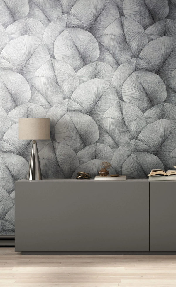 Marburg Kumano 34508 Large-scale gray fan-shaped leaf wallpaper behind a gray console with a metallic table lamp, a decorative knot, a small glass vase, and an open book on a light wood floor.