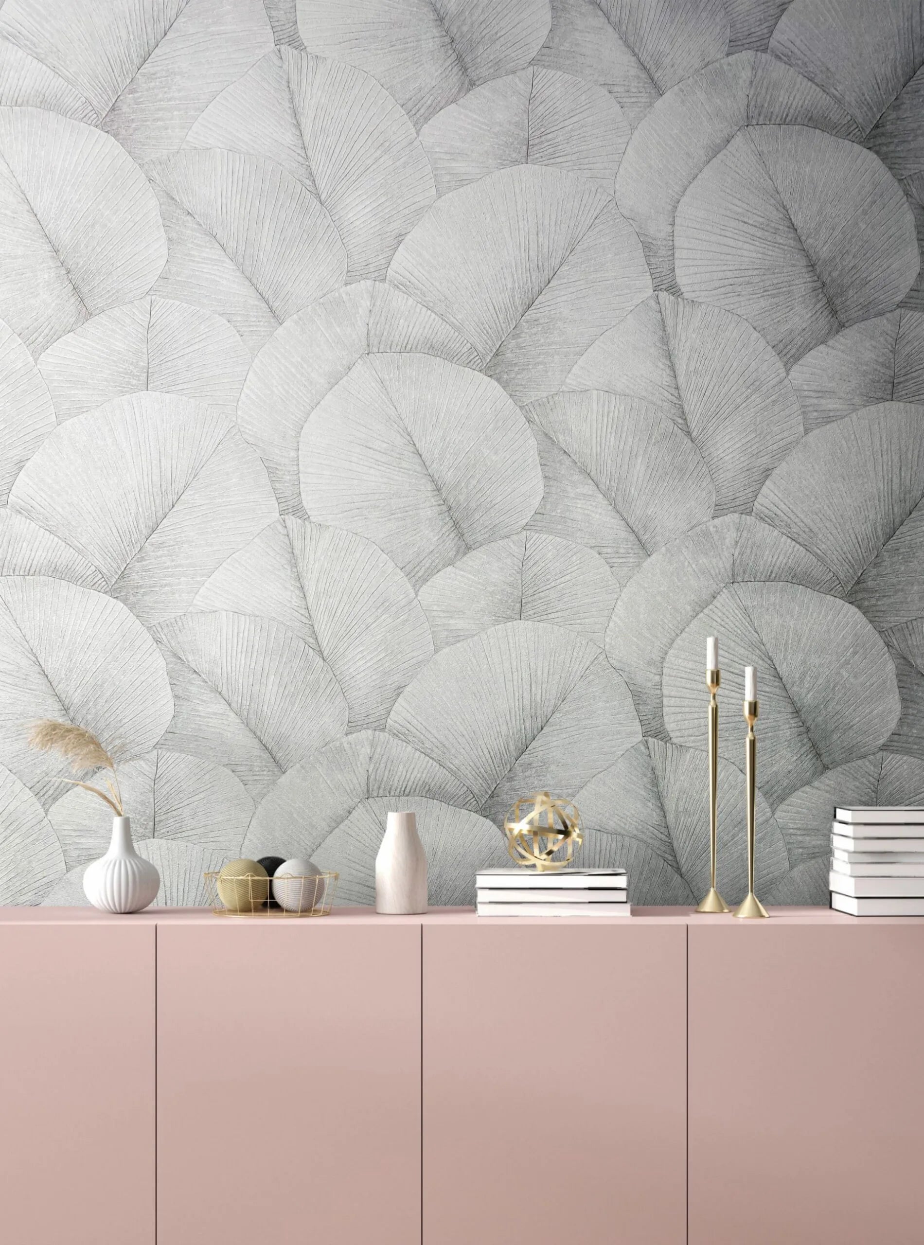 Marburg Kumano 34509 Gray overlapping fan-leaf wallpaper behind a pink sideboard styled with vases, a dried grass stem, a wire basket with decorative balls, stacked books, a gold orb sculpture, and brass candlesticks with white candles.