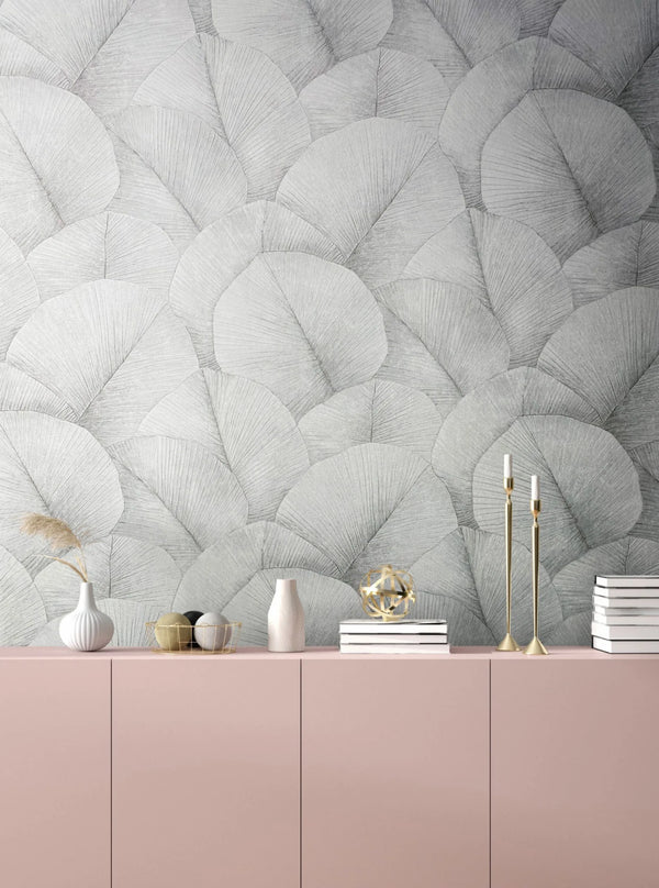 Marburg Kumano 34509 Gray overlapping fan-leaf wallpaper behind a pink sideboard styled with vases, a dried grass stem, a wire basket with decorative balls, stacked books, a gold orb sculpture, and brass candlesticks with white candles.