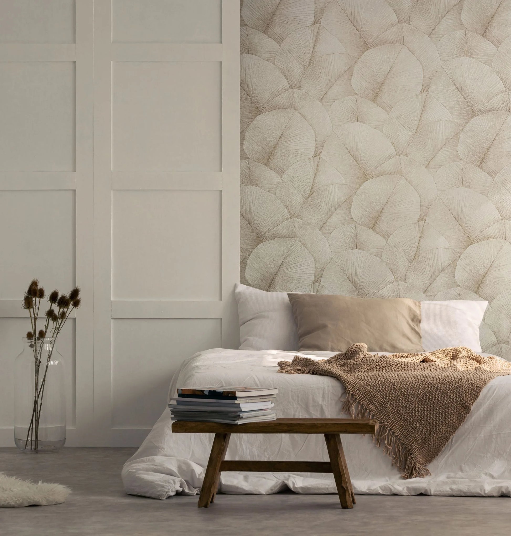 Marburg Kumano 34510 Bedroom scene with a feature wall of beige leaf-patterned wallpaper behind a low bed with pillows and a knitted throw, a wooden bench with stacked books, and a glass vase of dried stems.