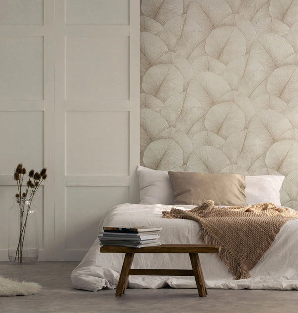 Marburg Kumano 34510 Bedroom scene with a feature wall of beige leaf-patterned wallpaper behind a low bed with pillows and a knitted throw, a wooden bench with stacked books, and a glass vase of dried stems.