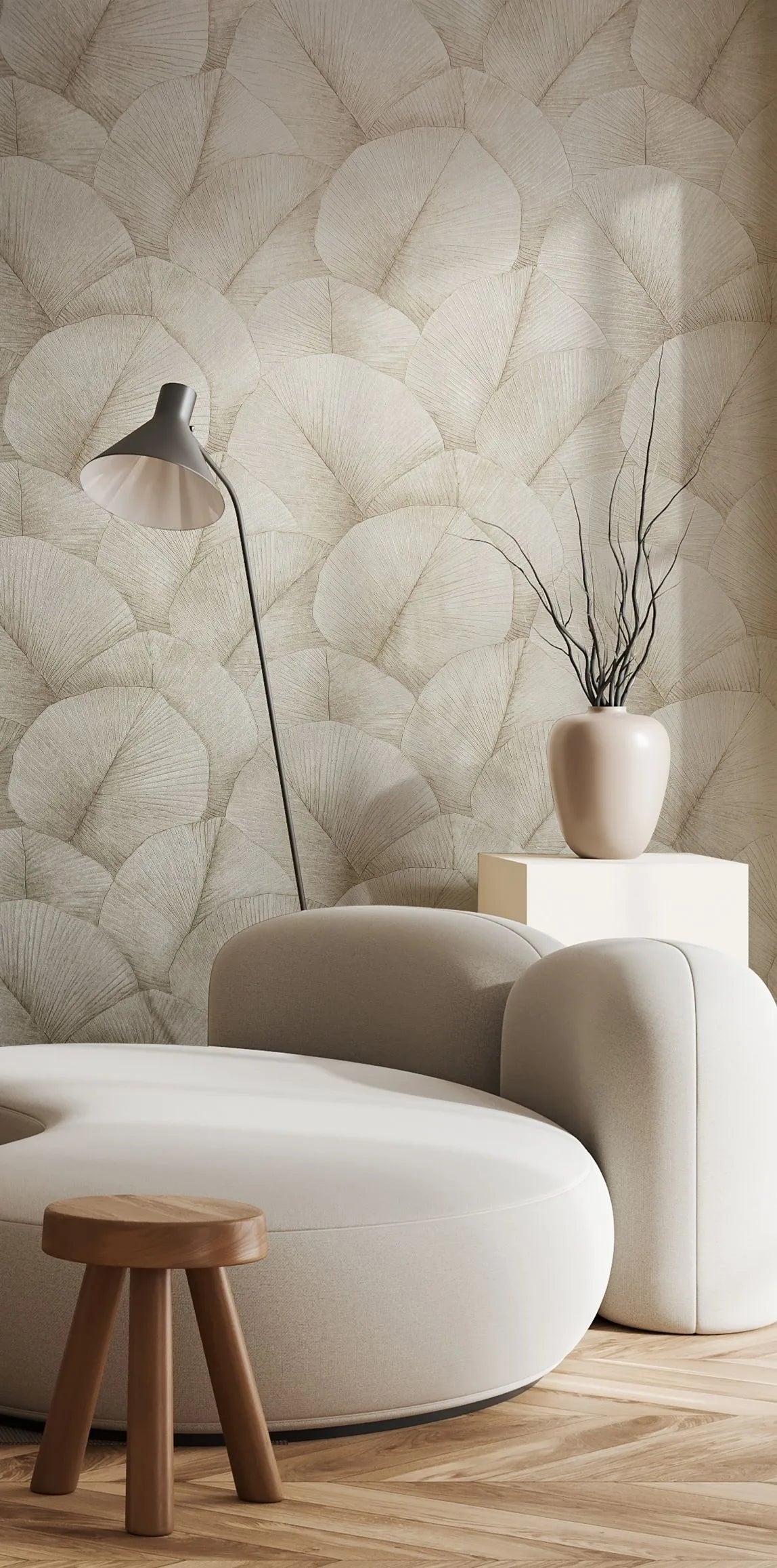 Marburg Kumano 34510 Living room scene with beige wallpaper of large overlapping fan-shaped leaves, shown behind a curved cream sofa, small wooden stool, floor lamp, and a ceramic vase with branches on a pedestal.