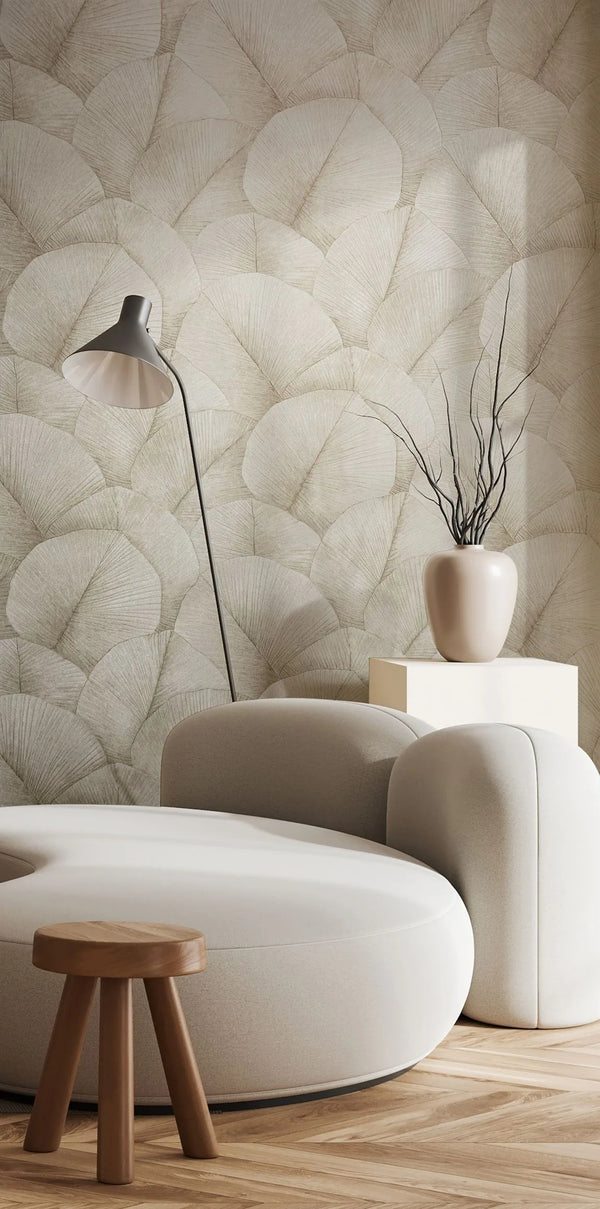 Marburg Kumano 34510 Living room scene with beige wallpaper of large overlapping fan-shaped leaves, shown behind a curved cream sofa, small wooden stool, floor lamp, and a ceramic vase with branches on a pedestal.