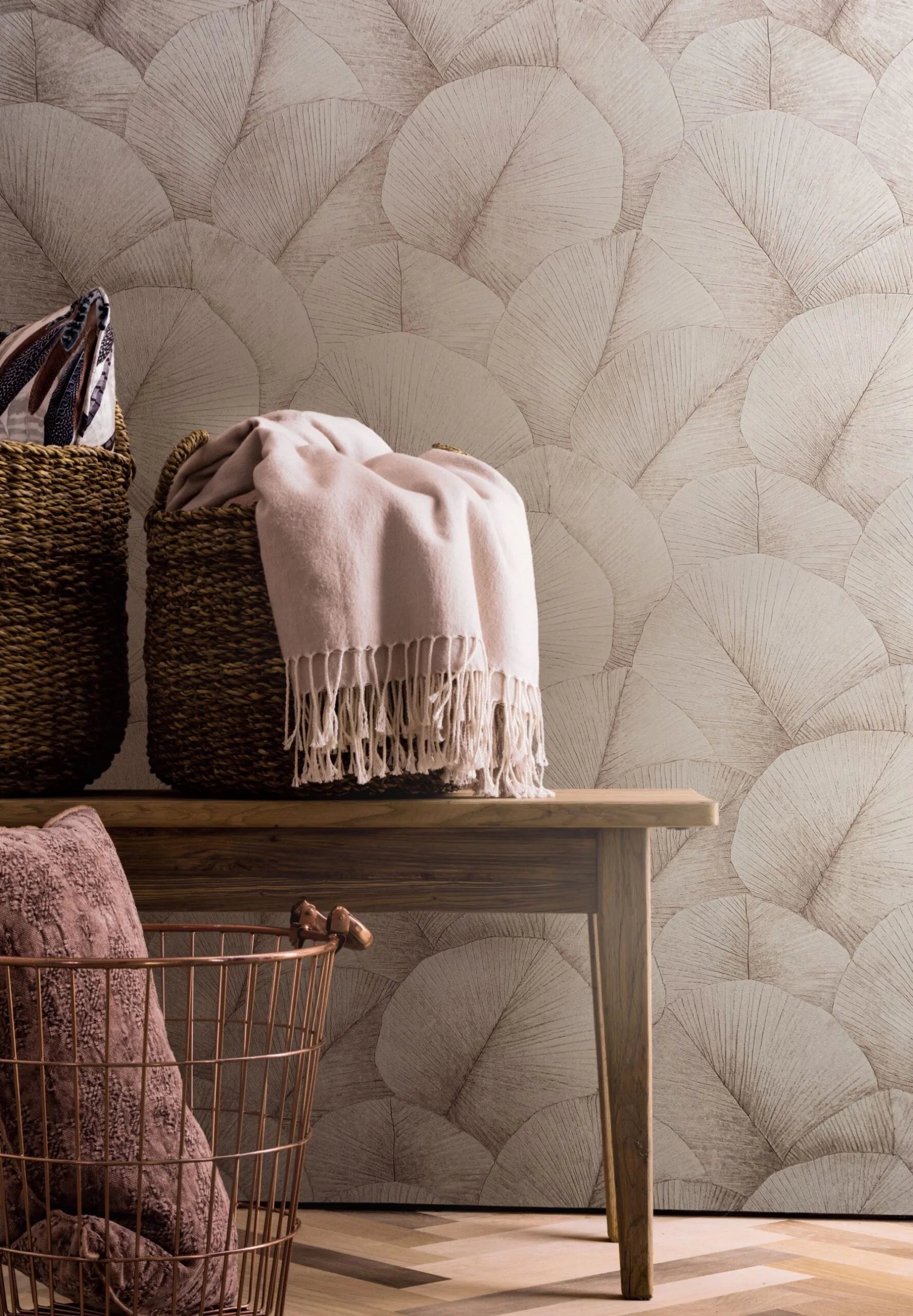Marburg Kumano 34511 Wallpaper with large overlapping fan-leaf pattern behind a wooden bench holding woven baskets and a fringed throw, with a wire basket and cushion on a herringbone wood floor.