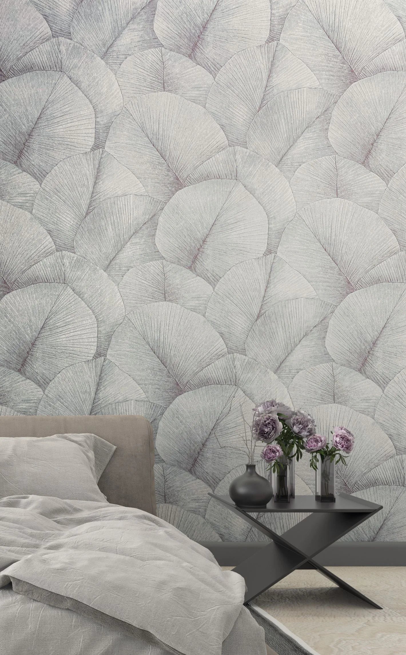 Marburg Kumano 34512 Bedroom scene featuring a large-scale leaf pattern wallpaper in light gray behind a bed with gray bedding and a black side table holding vases with purple flowers.