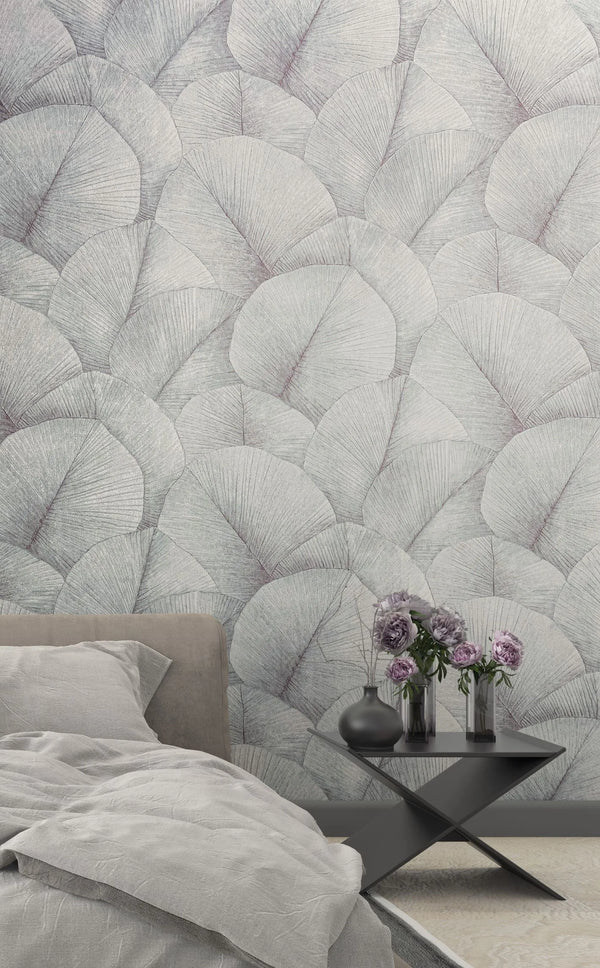 Marburg Kumano 34512 Bedroom scene featuring a large-scale leaf pattern wallpaper in light gray behind a bed with gray bedding and a black side table holding vases with purple flowers.