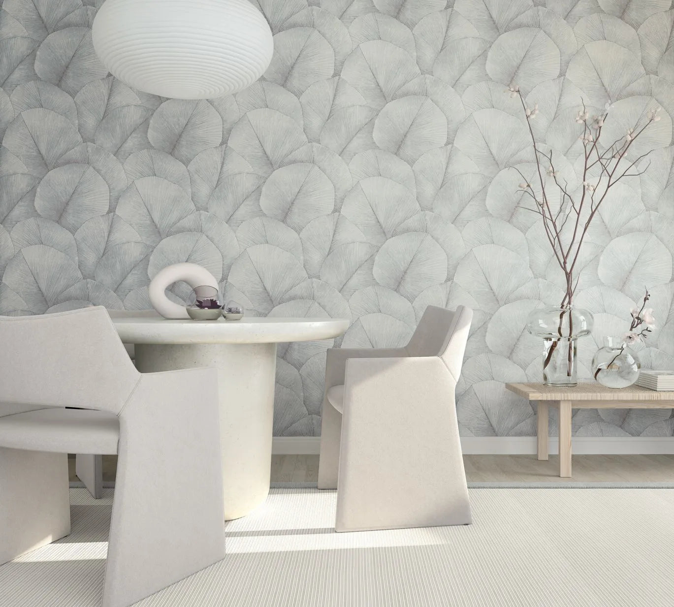 Marburg Kumano 34512 Room scene with a wall covered in soft gray overlapping fan‑leaf wallpaper behind a round light-toned dining table, sculptural armchairs, a pendant light, and a wooden bench holding clear glass vases with branches.