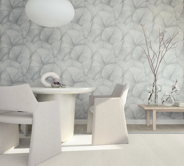 Marburg Kumano 34512 Room scene with a wall covered in soft gray overlapping fan‑leaf wallpaper behind a round light-toned dining table, sculptural armchairs, a pendant light, and a wooden bench holding clear glass vases with branches.