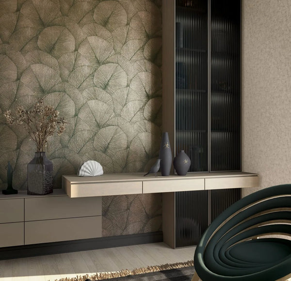 Marburg Kumano 34513 Leaf-pattern wallpaper in green and gold shown as a feature wall behind a floating sideboard with decorative vases, a shell object, dried branches in a glass vase, and a ribbed-glass cabinet, with a sculptural dark green chair in the foreground.
