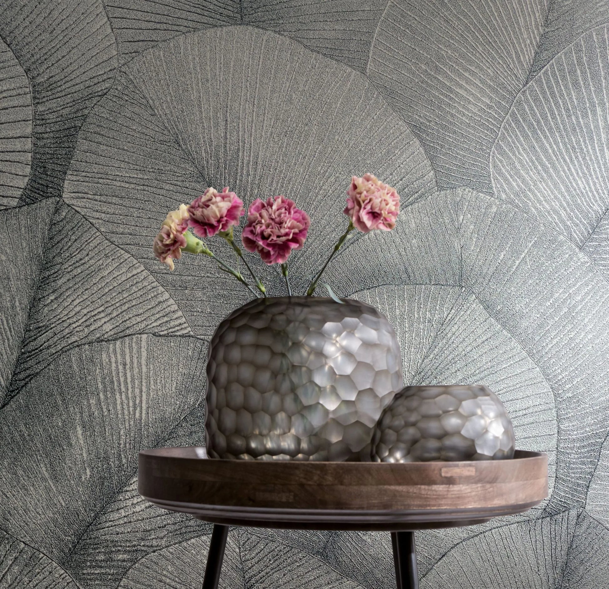 Marburg Kumano 34514 Silver wallpaper with large overlapping fan-like leaf veins. In front, a wooden tray on a small table holds two textured silver-gray glass vessels, one with pink flowers.