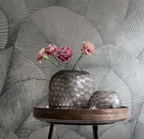Marburg Kumano 34514 Silver wallpaper with large overlapping fan-like leaf veins. In front, a wooden tray on a small table holds two textured silver-gray glass vessels, one with pink flowers.