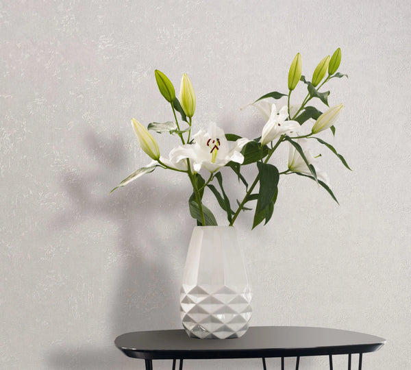 Marburg Kumano 34517 Light textured wallpaper behind a black table with a white faceted vase holding white lilies and green buds.