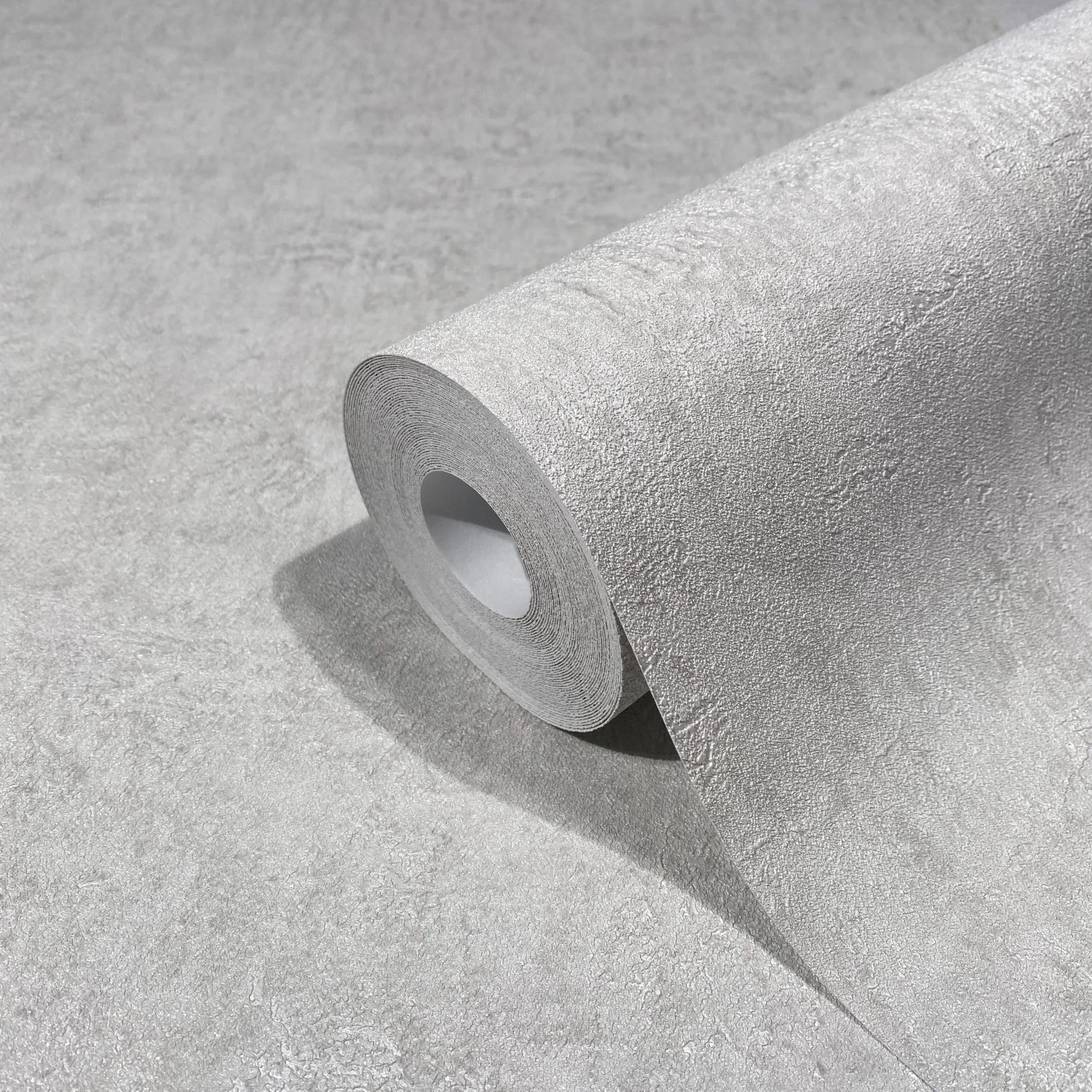 Marburg Kumano 34518 Close-up of a gray textured wallpaper roll resting on a matching unrolled sheet, showing a subtle plaster-like texture.