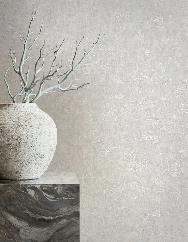 Marburg Kumano 34518 Textured light gray wallpaper with a subtle plaster effect behind a rough ceramic vase holding bare branches on a stone plinth.