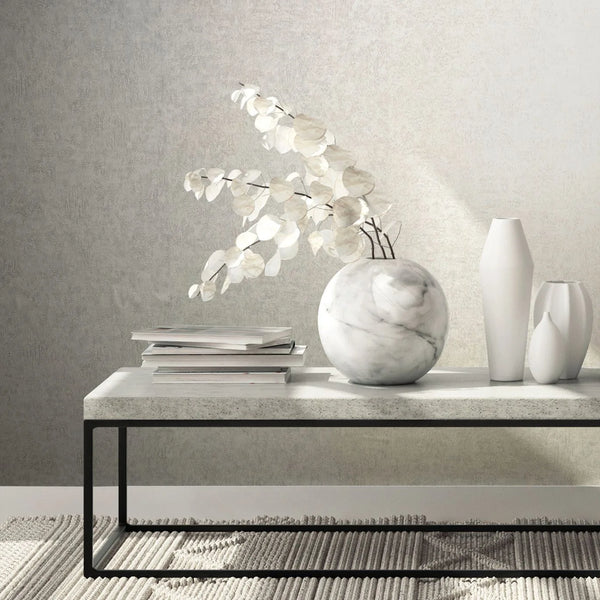 Marburg Kumano 34519 Gray textured wallpaper shown behind a minimalist table holding a round marble vase with branches, two white vases, and stacked books on a rug.