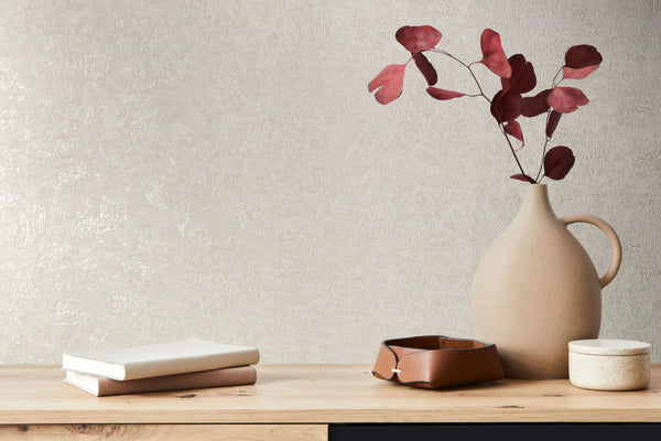 Marburg Kumano 34520 Beige textured wallpaper behind a wooden console styled with a matte ceramic jug holding red leaves, two stacked books, a leather tray, and a small lidded container.