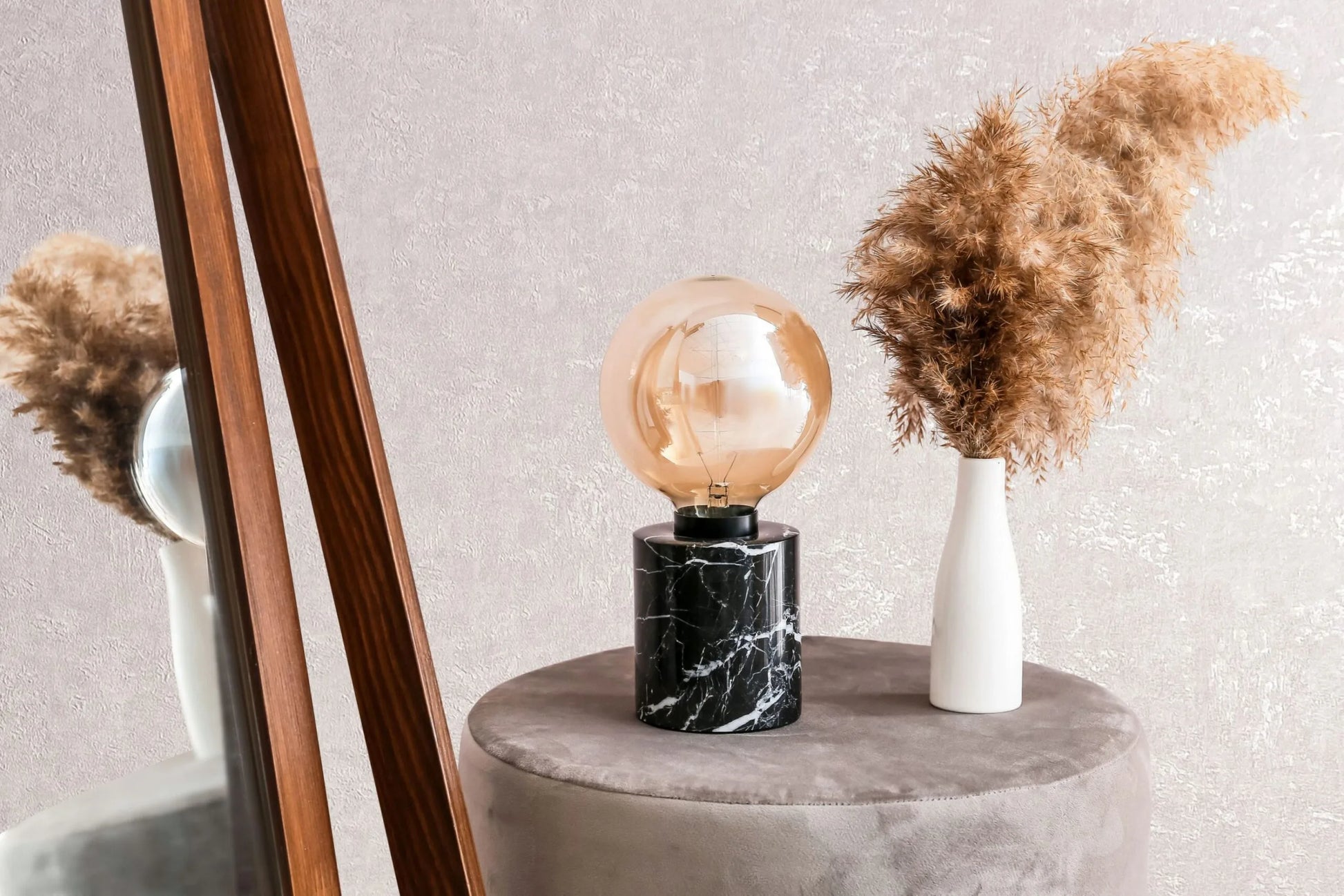 Marburg Kumano 34521 Soft pink textured wallpaper with a gentle sheen behind a black marble‑base lamp with an amber globe, a white vase of pampas on a round upholstered stool, and the edge of a leaning wooden mirror.