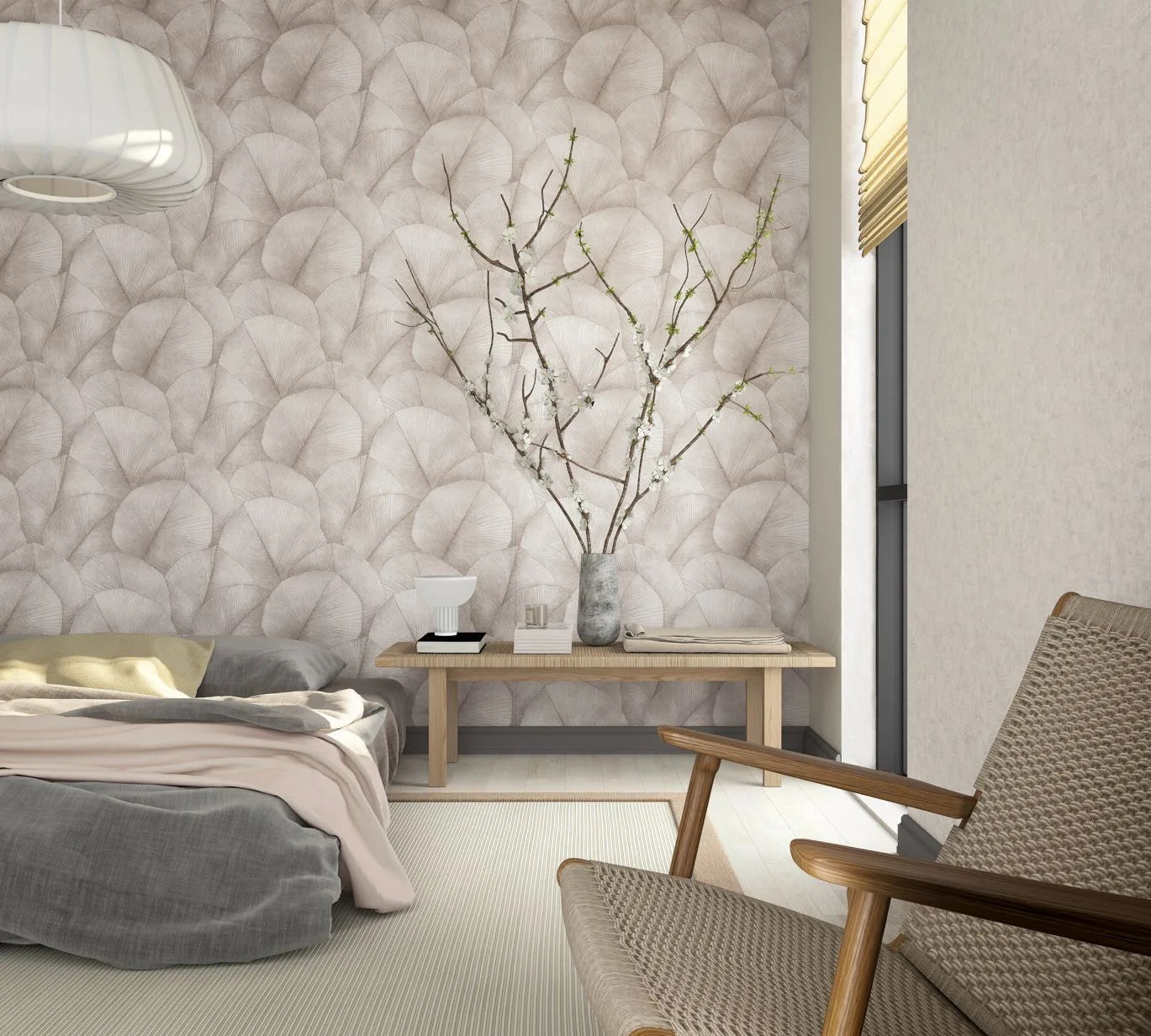 Marburg Kumano 34521 Bedroom scene with a feature wall covered in large overlapping leaf-pattern wallpaper in soft neutral tones, shown with a low bed, wooden bench holding a vase of blossoming branches and a small lamp, a woven lounge chair, and a window with a blind.