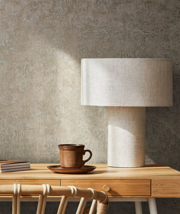 Marburg Kumano 34522 Textured plaster-look taupe wallpaper behind a wooden desk with a fabric-shaded table lamp, a brown cup and saucer, stacked notebooks, and a woven chair.