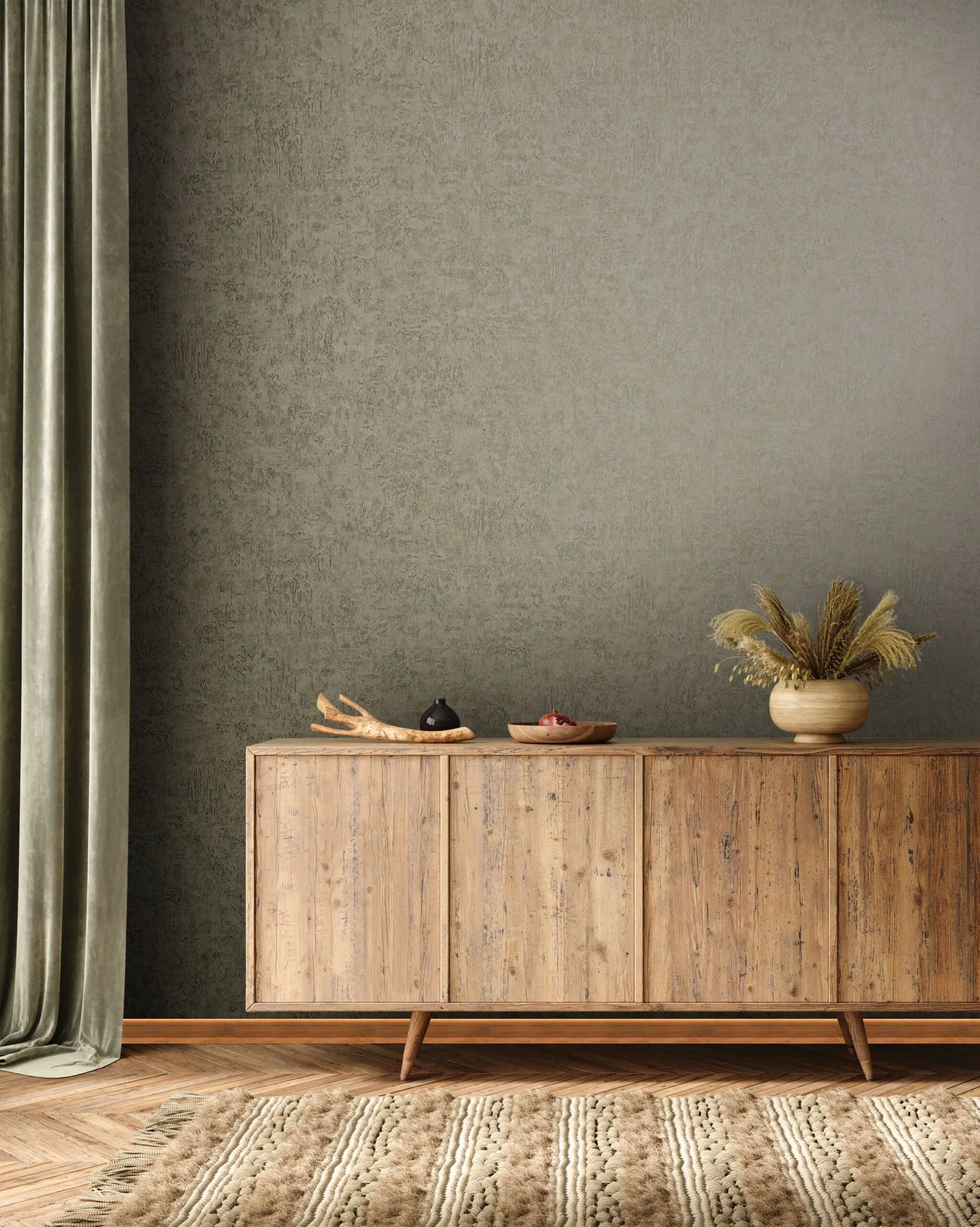 Marburg Kumano 34522 Olive-green mottled textured wallpaper behind a wooden sideboard with tabletop decor, a draped curtain at left, a pot with dried grasses, and a textured rug on a herringbone floor.