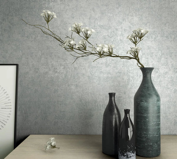 Marburg Kumano 34523 Blue-gray mottled wallpaper with a soft plaster effect behind three vases and a long flowering branch on a light wood tabletop, with a framed print partially visible at left.