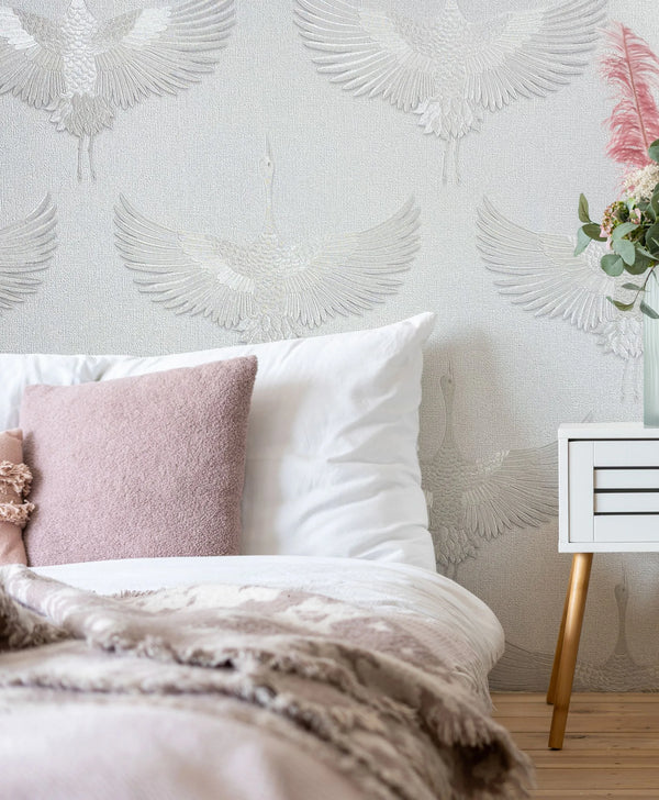 Marburg Kumano 34528 Tonal crane pattern wallpaper behind a bed with white bedding and a blush cushion, next to a white nightstand holding a vase of flowers.