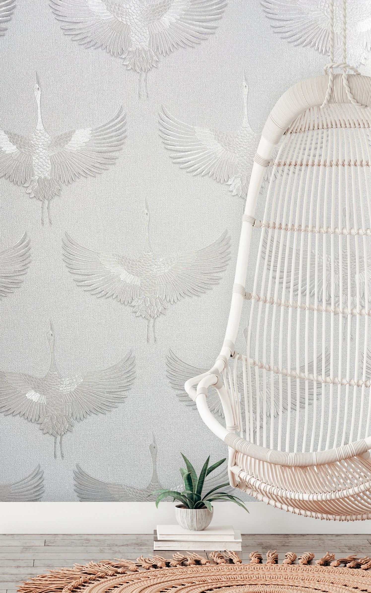 Marburg Kumano 34528 Light-colored wallpaper with a repeating crane-in-flight pattern. A rattan hanging chair is at right with a small potted plant nearby, a woven round rug, and wood flooring visible.