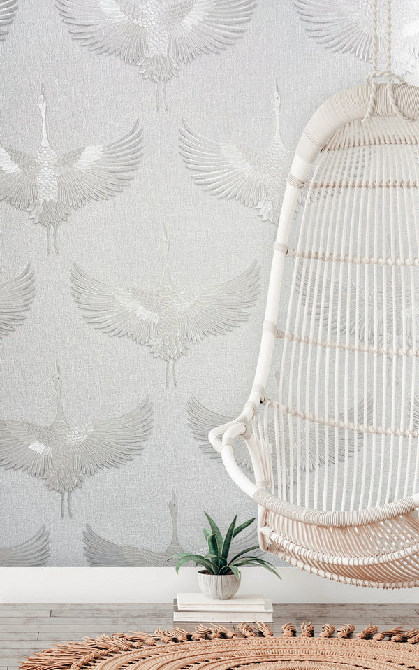 Marburg Kumano 34528 Light-colored wallpaper with a repeating crane-in-flight pattern. A rattan hanging chair is at right with a small potted plant nearby, a woven round rug, and wood flooring visible.