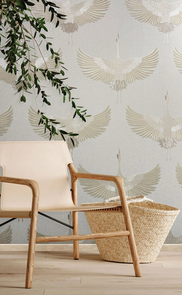 Marburg Kumano 34529 Textured wallpaper with repeating beige cranes on a light background, styled with a wood-framed lounge chair, a woven basket, and a hanging leafy branch on a wood floor.