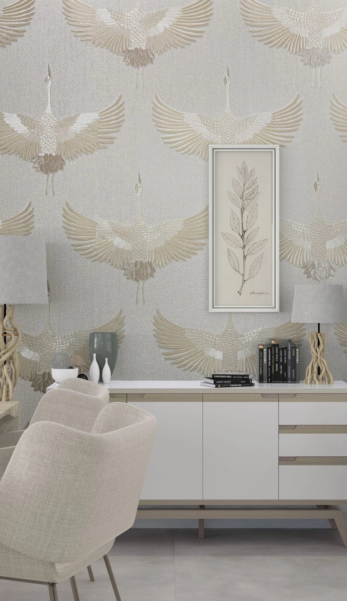 Marburg Kumano 34529 Room scene showing light textured wallpaper with large beige and white cranes in flight. In front is a white sideboard with books, decorative vases, and two sculptural table lamps, a framed botanical print on the wall, and a beige upholstered chair.