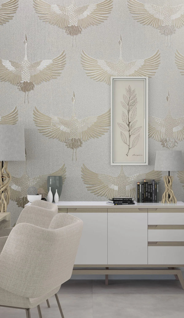 Marburg Kumano 34529 Room scene showing light textured wallpaper with large beige and white cranes in flight. In front is a white sideboard with books, decorative vases, and two sculptural table lamps, a framed botanical print on the wall, and a beige upholstered chair.
