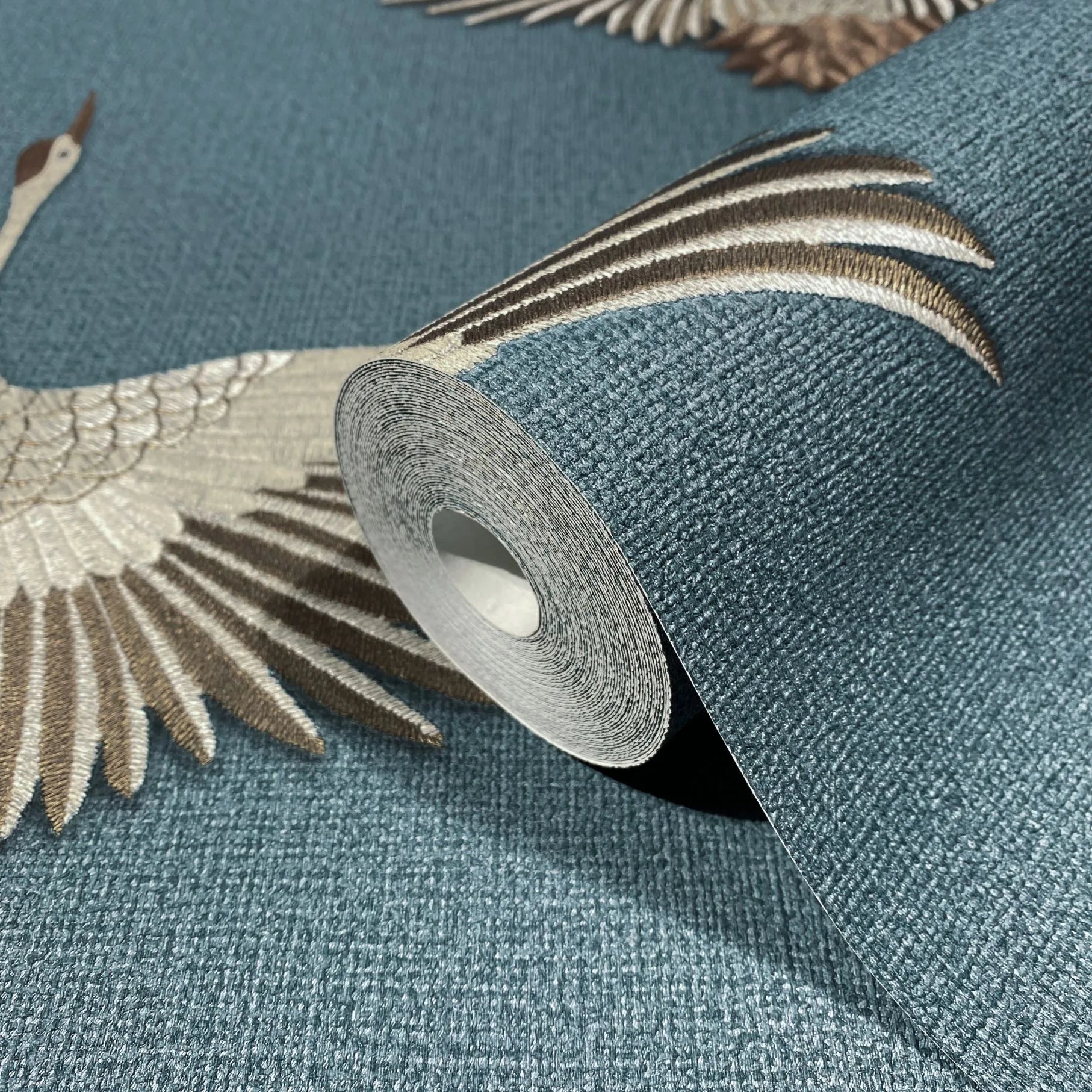 Marburg Kumano 34531 Close-up of a wallpaper roll resting on matching textured blue wallpaper featuring flying crane illustrations in beige and brown.