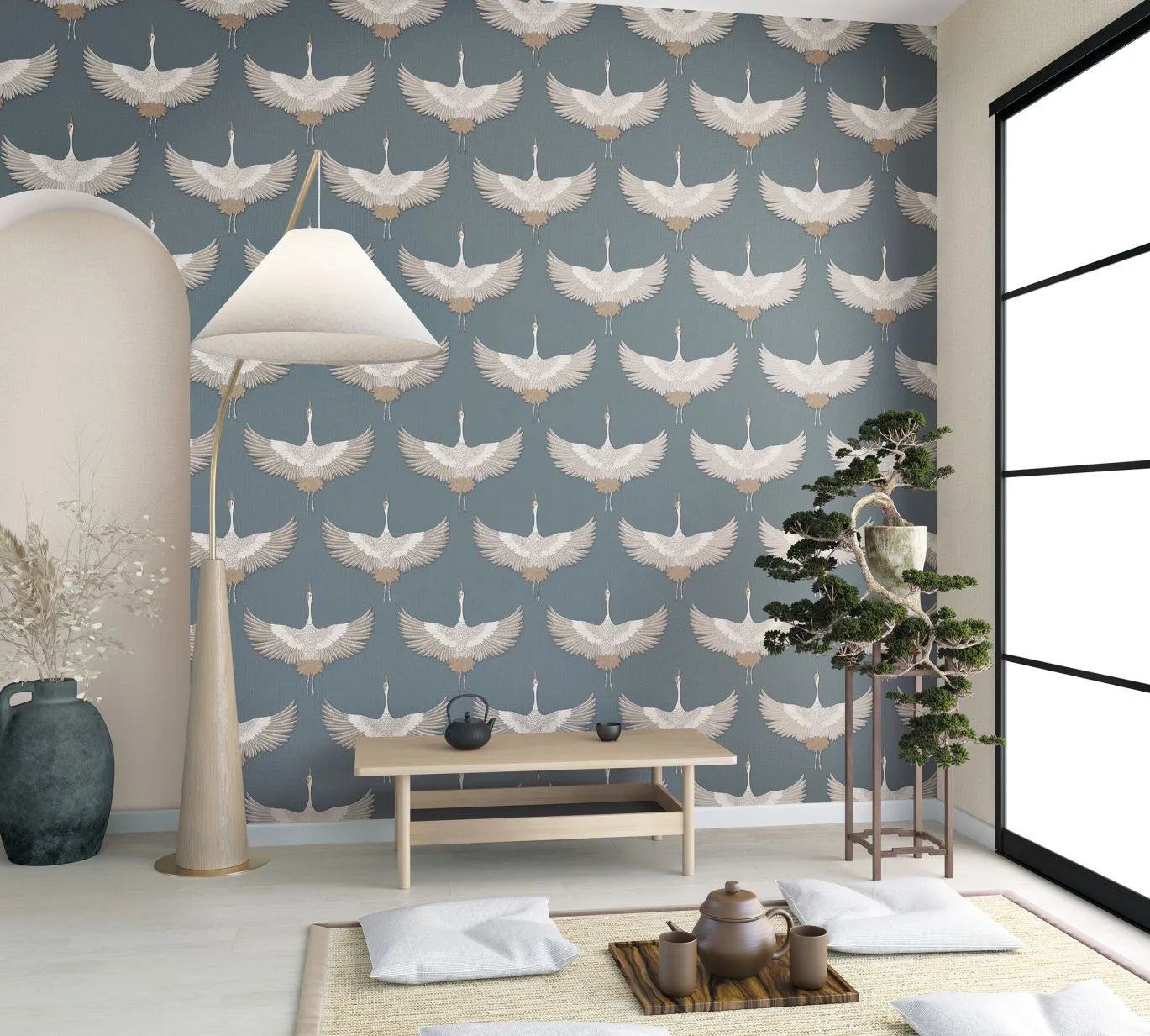 Marburg Kumano 34531 Blue wallpaper with a repeating white crane motif shown on a feature wall behind a light wood bench. A floor lamp, large vase with dried stems, floor cushions with a mat and tea set, a bonsai on a stand, and a large grid-frame window are visible.