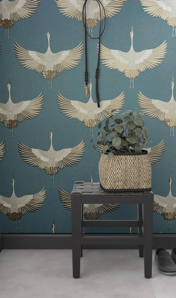 Marburg Kumano 34531 Blue wallpaper with a repeating beige crane pattern behind a black stool topped with a woven planter and leafy plant; a strap hangs on the wall and a pair of shoes is on the floor.