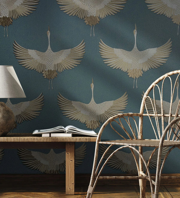 Marburg Kumano 34531 Blue wallpaper with large beige cranes in flight behind a rattan chair, a wooden bench with an open book, and a table lamp.