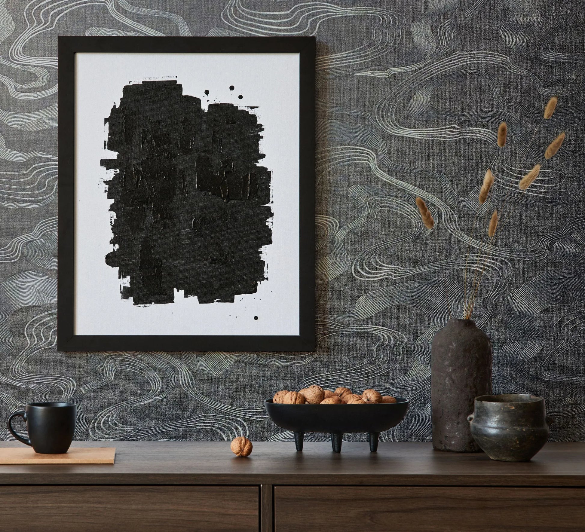 Marburg Kumano 34537 Dark wallpaper with fine, wavy contour lines forms the backdrop for a styled console scene with a framed abstract artwork, a mug, a pedestal bowl of nuts, a tall vase with dried stems, and a small ceramic pot.