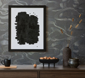 Marburg Kumano 34537 Dark wallpaper with fine, wavy contour lines forms the backdrop for a styled console scene with a framed abstract artwork, a mug, a pedestal bowl of nuts, a tall vase with dried stems, and a small ceramic pot.