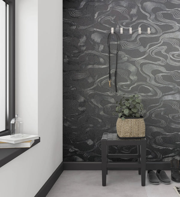 Marburg Kumano 34537 Black wallpaper with flowing silver contour lines in an entryway, shown behind a dark bench with a woven planter, wall hooks with a lanyard, a window ledge with books and a glass cloche, and shoes on the floor.