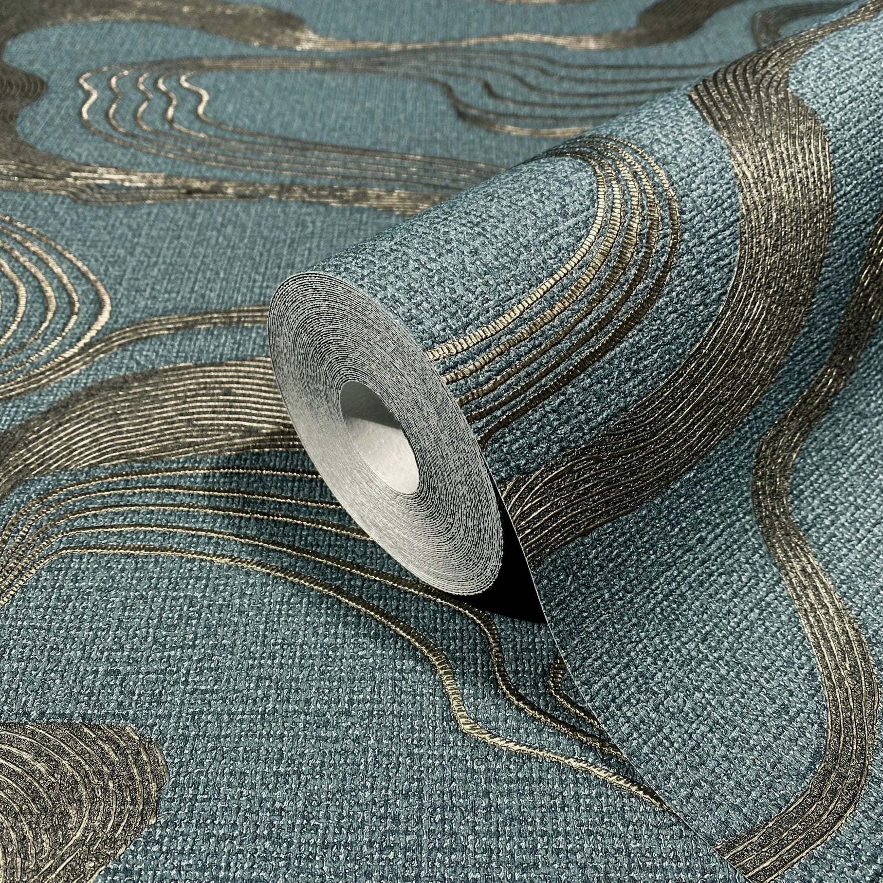 Marburg Kumano 34538 Close-up of a blue textured wallpaper roll with raised metallic gold curved lines, shown partially unrolled on matching wallpaper.