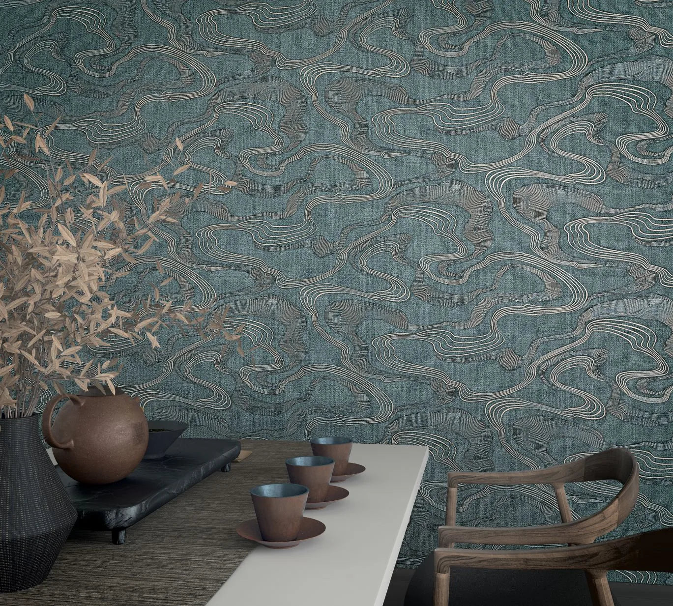 Marburg Kumano 34538 Blue wallpaper with flowing contour-like lines in gray and light tones, shown behind a tabletop with cups and saucers, a ceramic jug on a tray, a vase with branches, and a wooden chair.