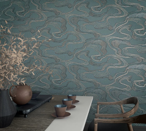 Marburg Kumano 34538 Blue wallpaper with flowing contour-like lines in gray and light tones, shown behind a tabletop with cups and saucers, a ceramic jug on a tray, a vase with branches, and a wooden chair.