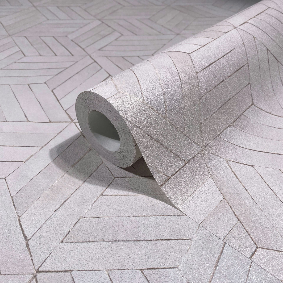 Marburg Leyla 81699 A close-up of a wallpaper roll resting on matching unrolled wallpaper with a light gray, textured geometric chevron pattern that forms radiating shapes.