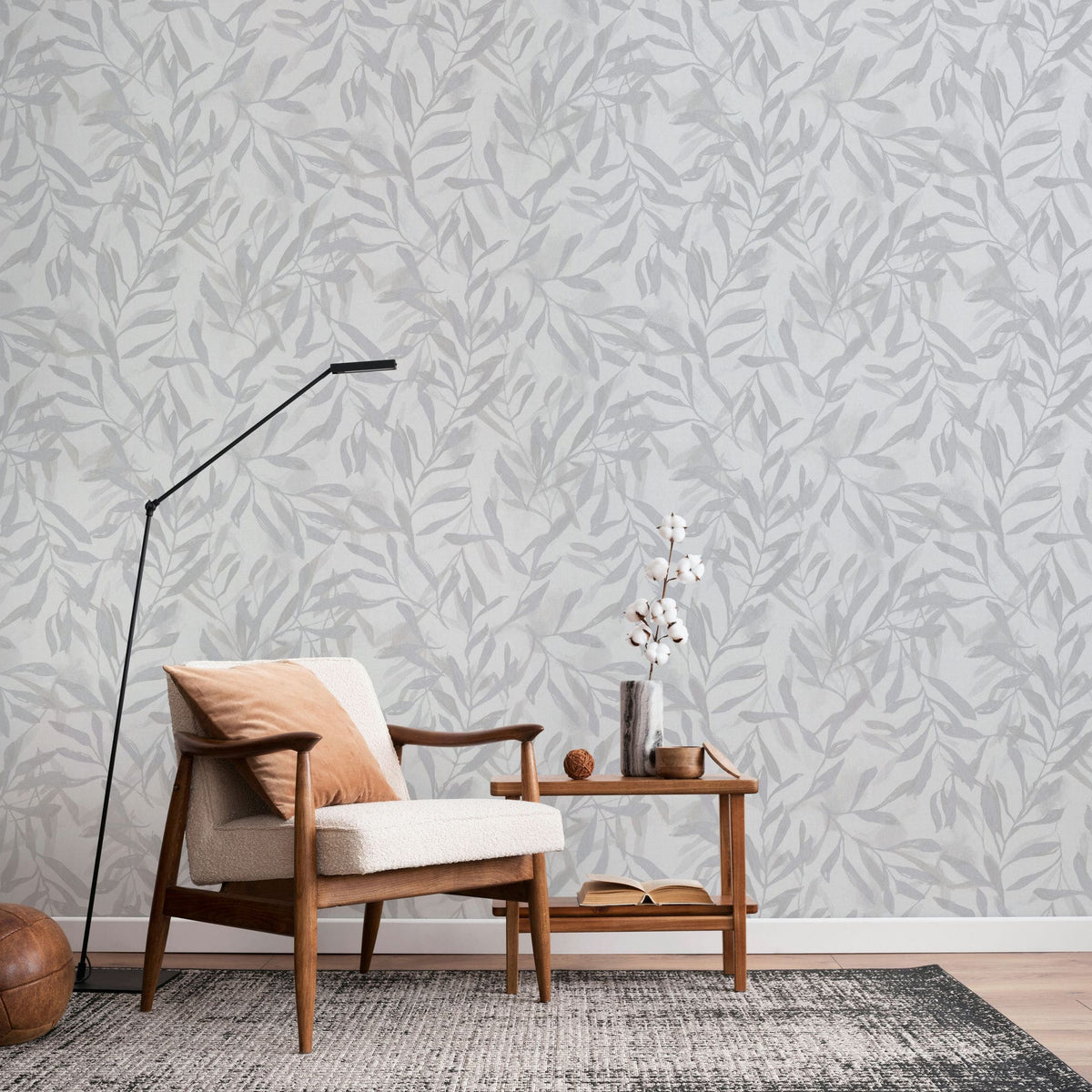 Marburg Leyla 82439 Light gray wallpaper with a tonal leaf pattern shown in a living room scene with a wooden armchair, black floor lamp, side table with a vase of white stems, and a rug.