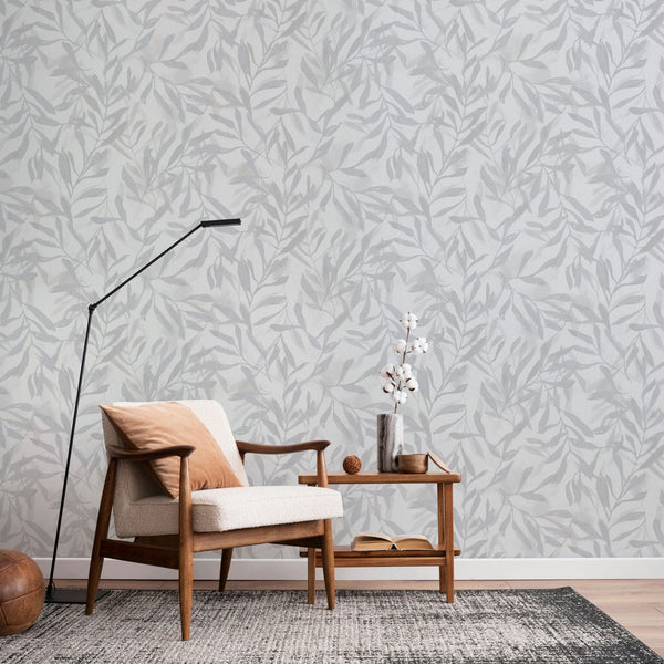 Marburg Leyla 82439 Light gray wallpaper with a tonal leaf pattern shown in a living room scene with a wooden armchair, black floor lamp, side table with a vase of white stems, and a rug.