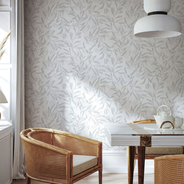Marburg Leyla 82439 Light gray and white leafy wallpaper behind a dining setup with rattan chairs, a white-topped table, a white pendant light, and a window with curtains; teapot and cups on the table.