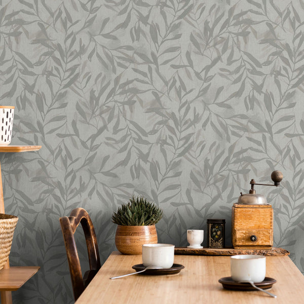 Marburg Leyla 82440 Tonal gray‑beige wallpaper with overlapping leafy branches behind a wooden table set with cups, saucers, spoons, a potted plant, and a manual coffee grinder.