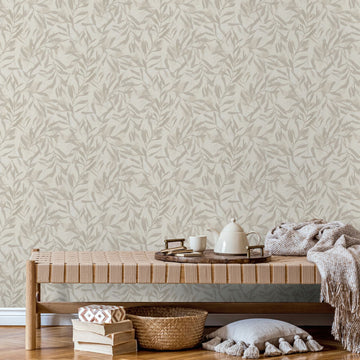 Marburg Leyla 82441 Beige wallpaper with a tone-on-tone leafy pattern behind a woven bench with a wooden tray, teapot, cup, throw, stacked books, a woven basket, and a tasseled floor cushion.